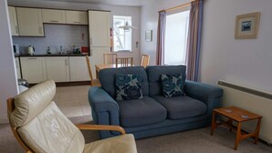 Un espacio habitable pequeño con un sofá, una silla, una mesita, una zona de cocina y una ventana con cortinas.
