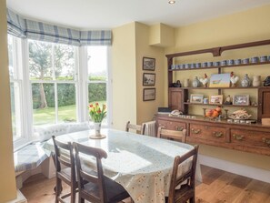 Un coin repas avec une table dressée pour deux personnes, une fenêtre en saillie agrémentée de rideaux et un meuble en bois orné d’objets décoratifs.