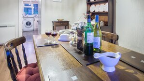 A dining table with wine glasses, bottles, and a white gravy boat. A wooden chair with a cushion is visible.
