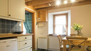 Een keuken met houten kastjes, een Beko-koelkast, een eettafel met stoelen, een raam met zonwering, en een vaas met bloemen.