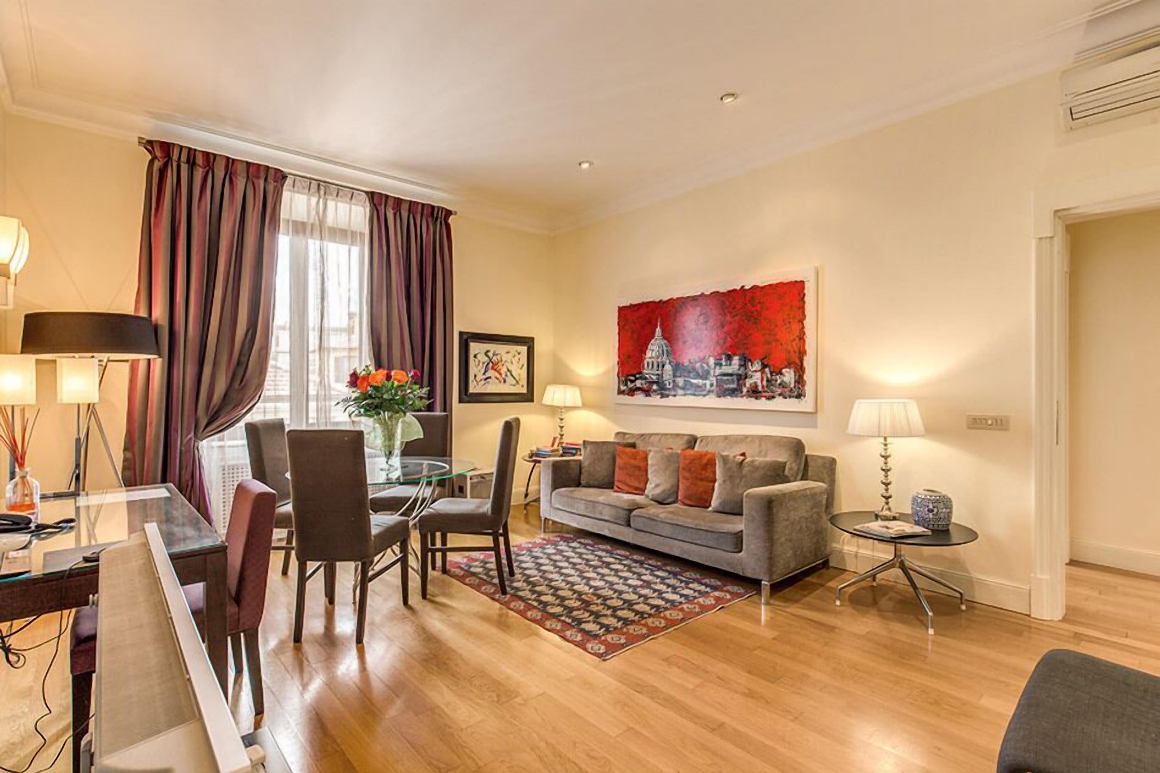 A modern living room with a dining area, a grey sofa, a red and white painting, and a glass coffee table.