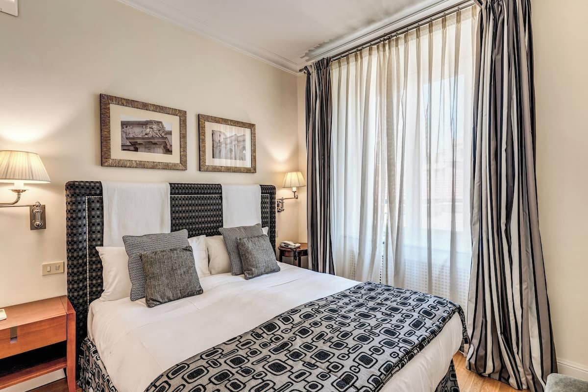 Una cama bien hecha con ropa de cama estampada, dos cuadros en la pared y un ventanal con cortinas.