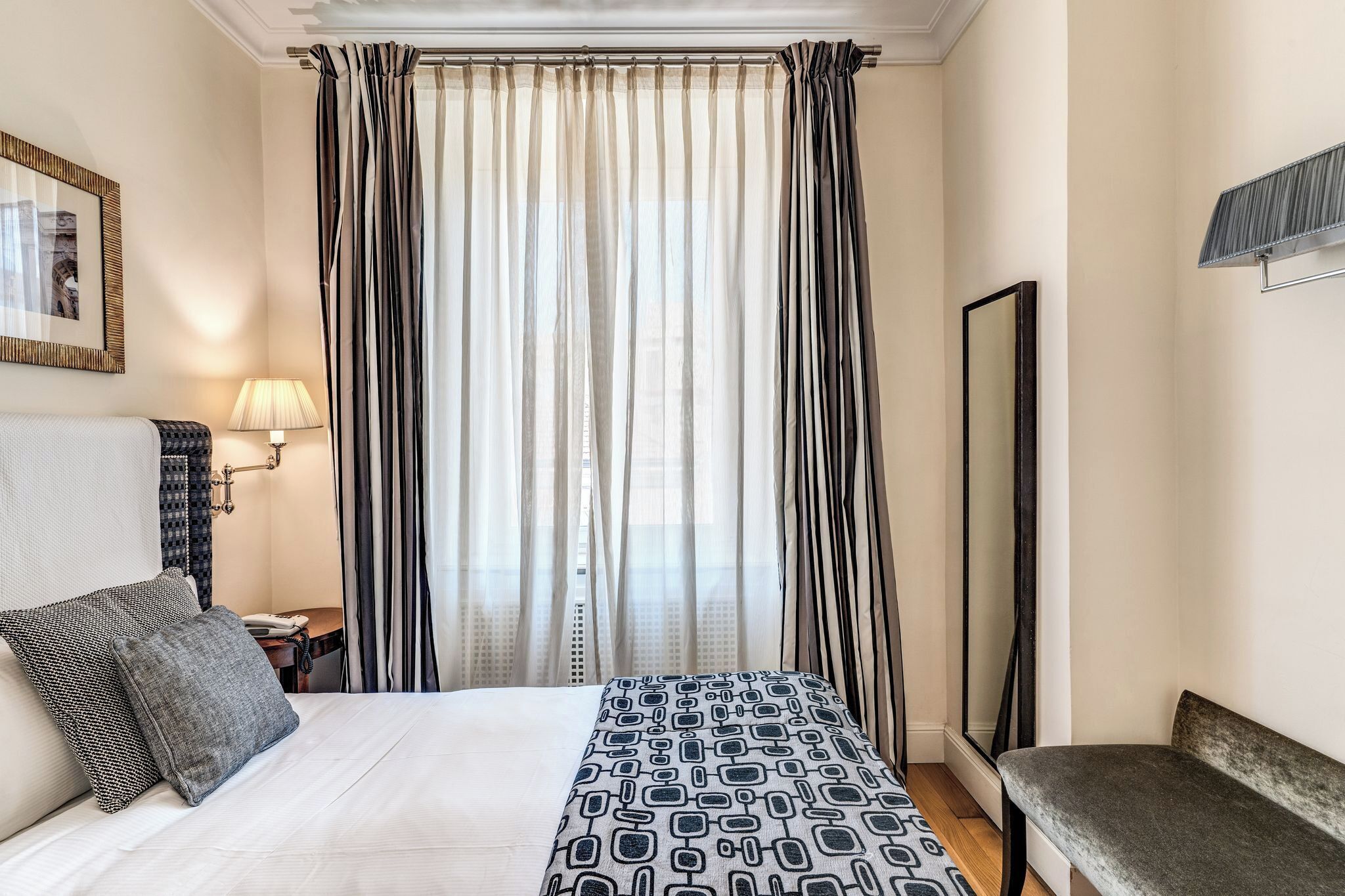 A hotel room with a bed, a bench, a nightstand with a lamp, and a large mirror.