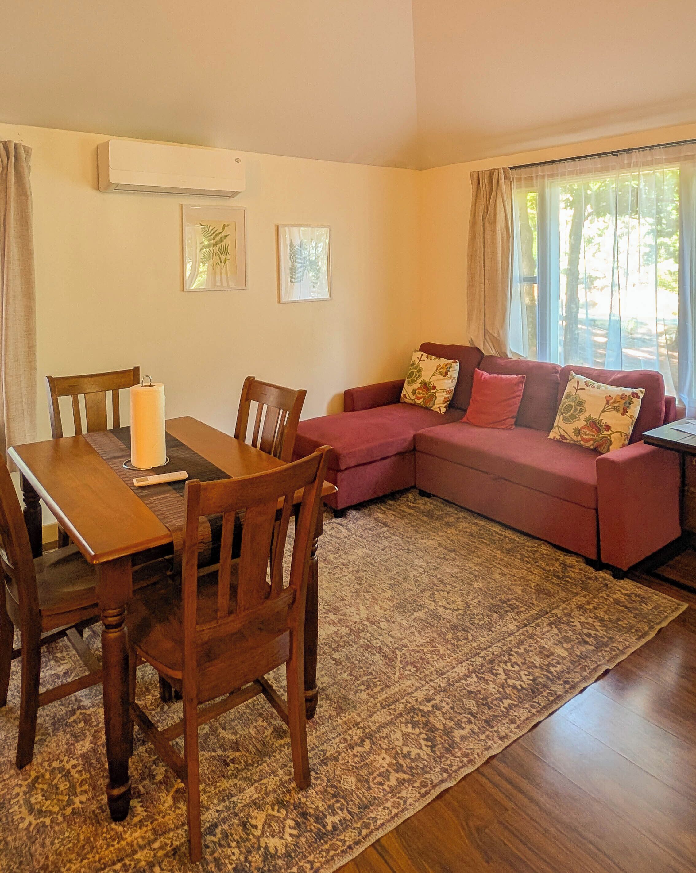 Uma sala de estar com uma mesa de jantar de madeira, duas cadeiras, um sofá vermelho com almofadas estampadas, um tapete estampado e uma janela com cortinas.