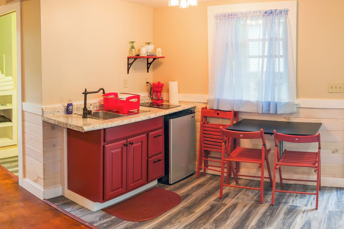 A compact kitchen with a sink, a coffee maker, and a small refrigerator. There is a window with sheer curtains and a wooden floor.