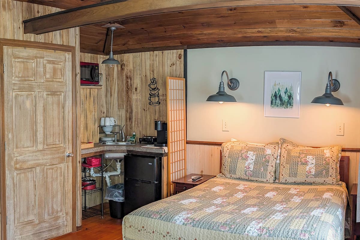 A bedroom with a large bed, wooden ceiling, a small kitchenette, and a wooden door.