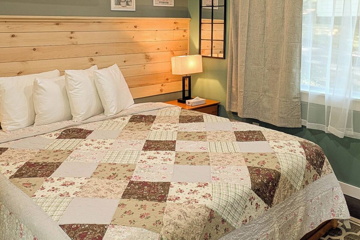 A bedroom with a wooden headboard, a quilted bedspread, a bedside lamp, and a window with curtains.