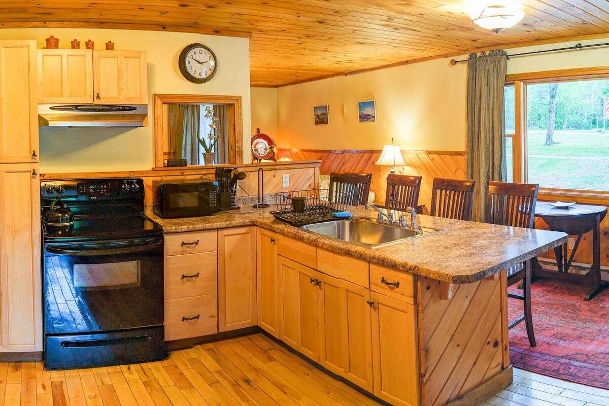 A kitchen with wooden cabinets, a stove, a microwave, and a sink. There is a clock on the wall and a window with curtains.