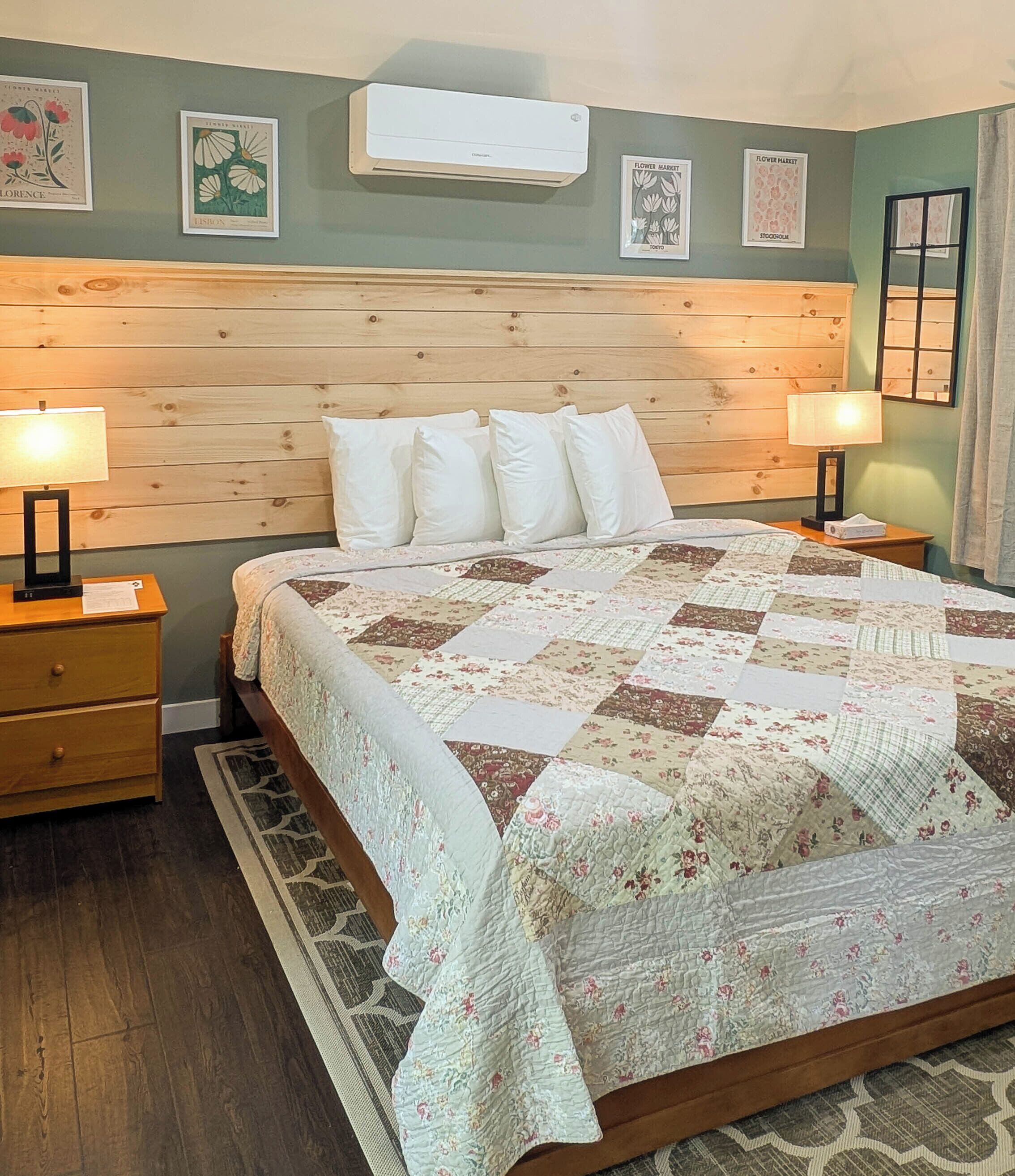 A bedroom with a wooden headboard, a quilted bedspread, two bedside tables with lamps, and framed floral artwork on the wall.