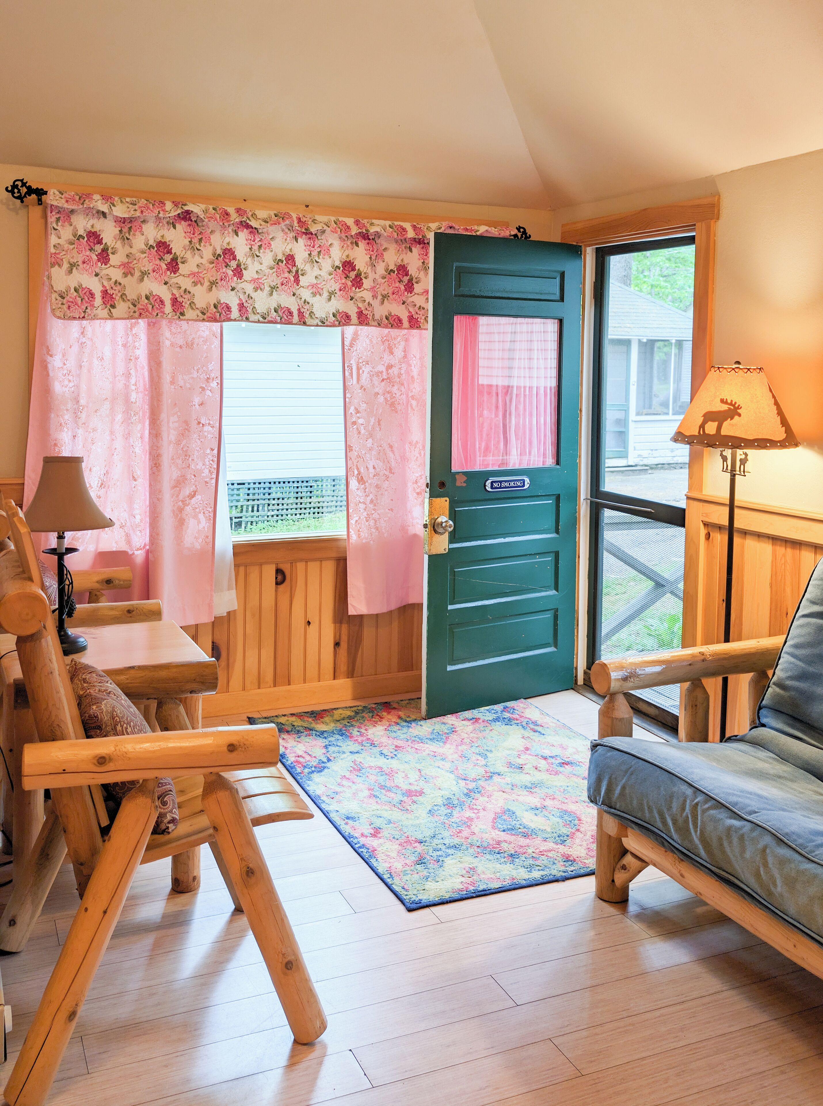 A cozy room with a wooden chair, a small table, a sofa, a colorful rug, and a green door with a 'No Smoking' sign.