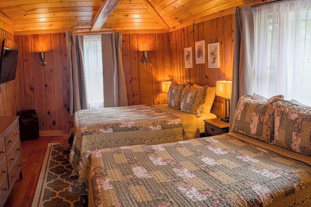 A cozy cabin-style bedroom with two beds, wooden walls, and a large window with curtains.