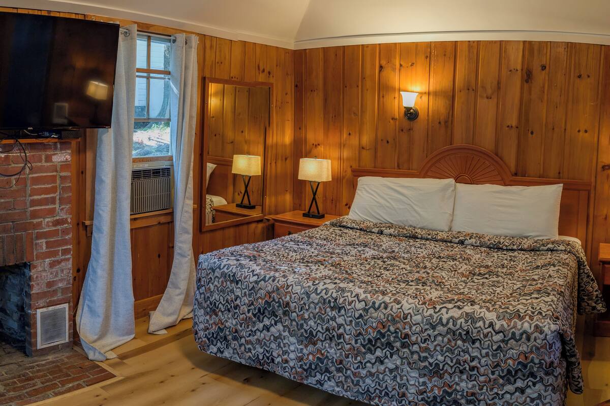 A bedroom with a wooden paneled wall, a bed with patterned bedding, a flat-screen TV, and a window with curtains.