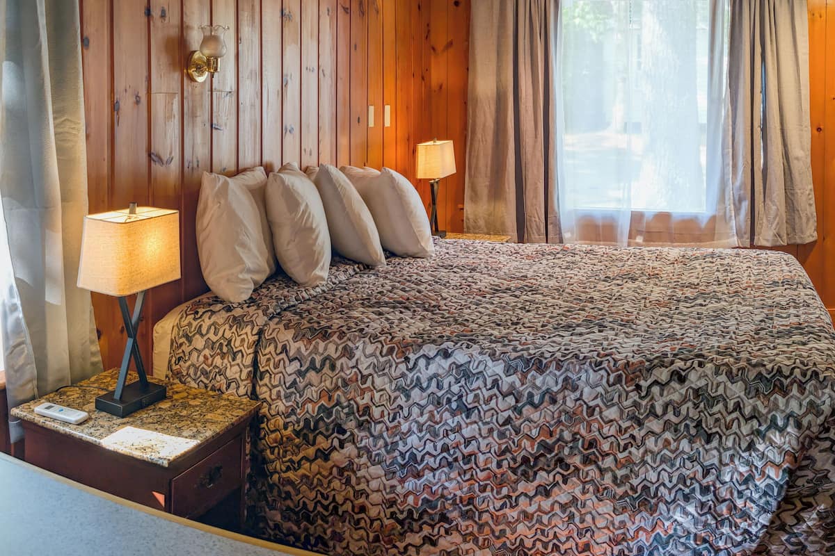 A wooden-paneled room with a bed, two bedside lamps, and a window with curtains.