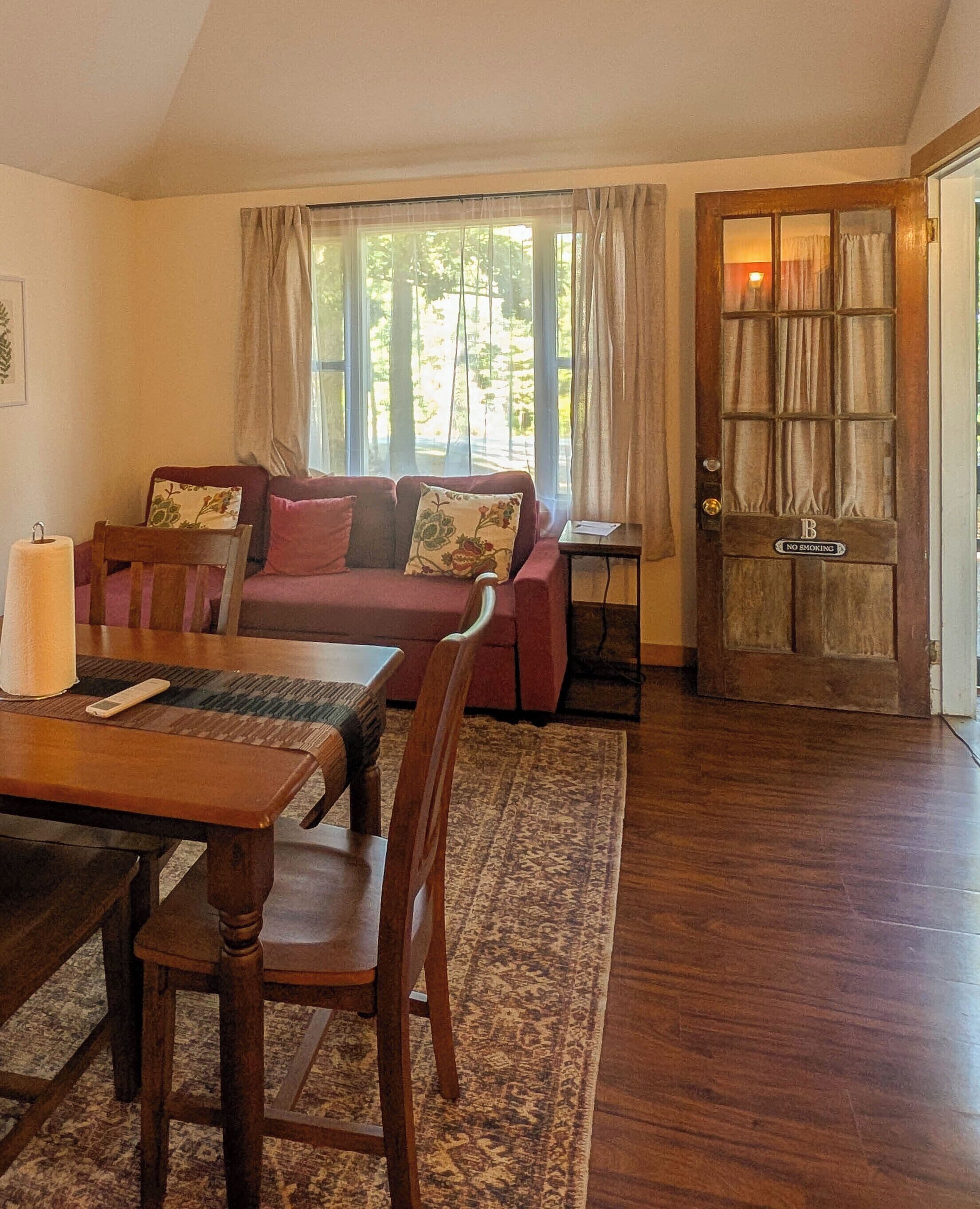 A cozy living room with a wooden dining table, a red sofa, and a patterned rug.