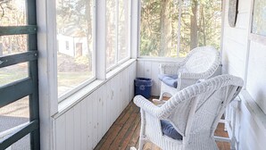 Een overdekte veranda met witte rotan schommelstoelen, houten vloeren en grote ramen met uitzicht op bomen en een gebouw.