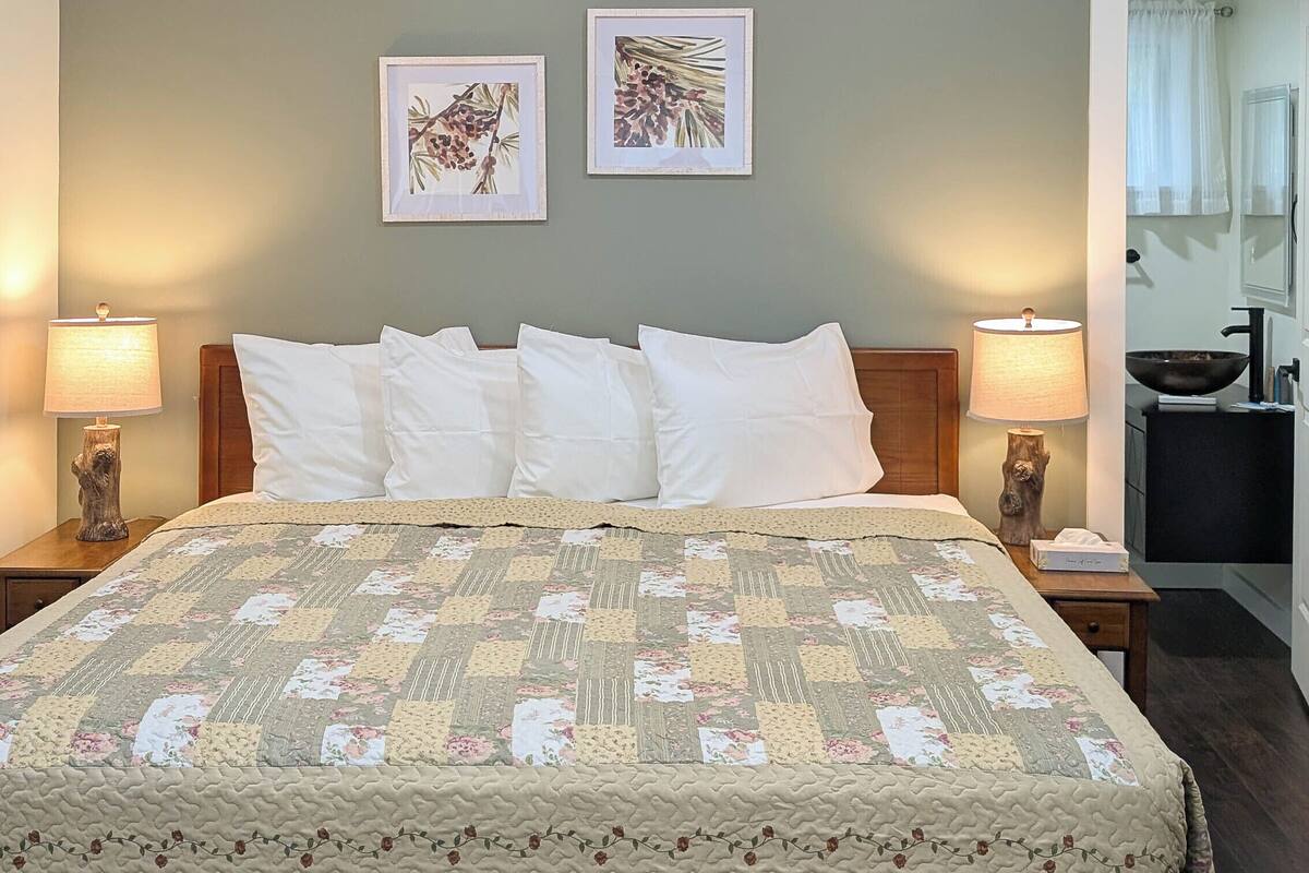 A neatly made bed with a floral quilt, two framed botanical prints on the wall, and a bathroom visible through an open door.