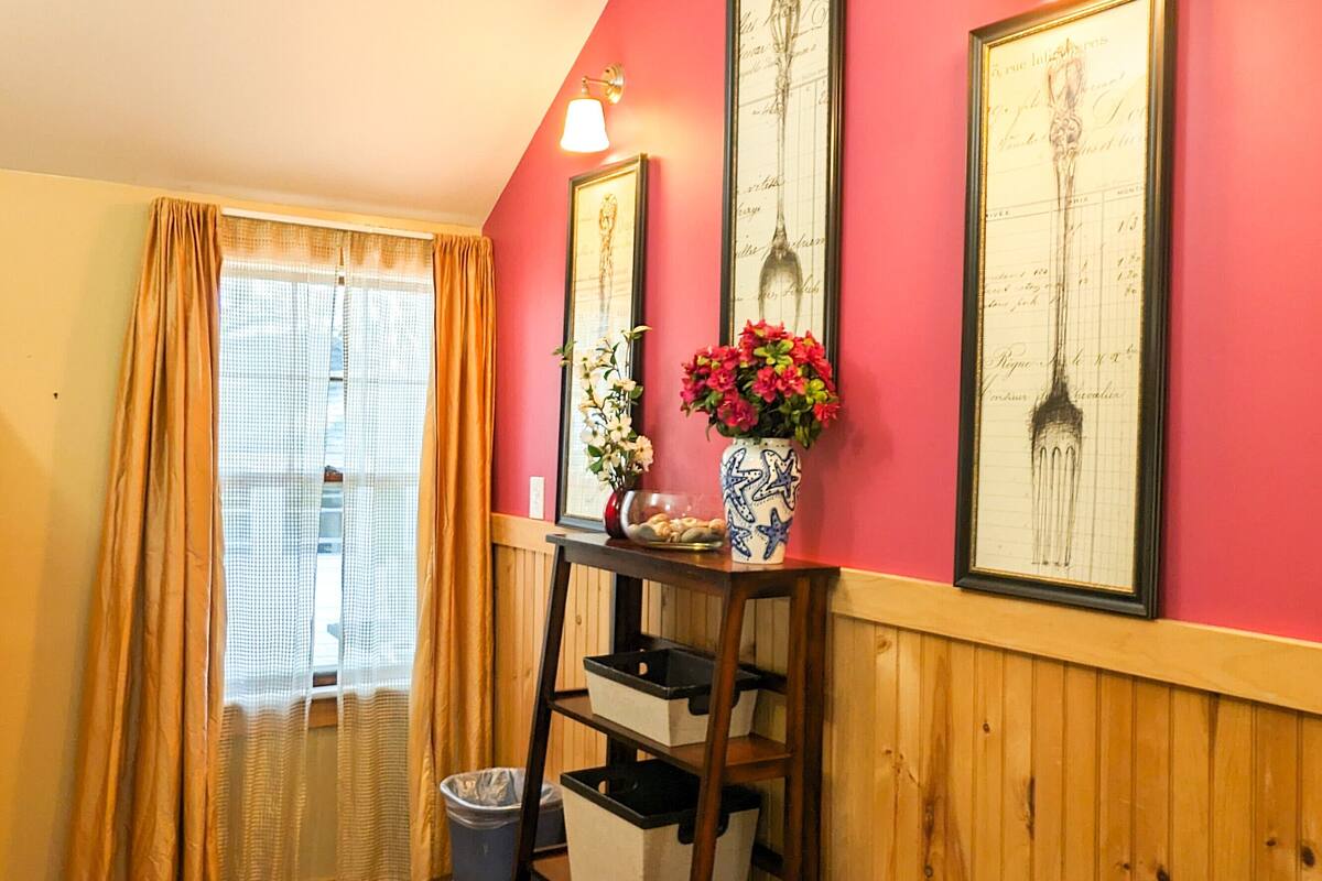 A room with wooden flooring, a window with curtains, a wall-mounted shelf with a vase of flowers, and framed pictures on the wall.