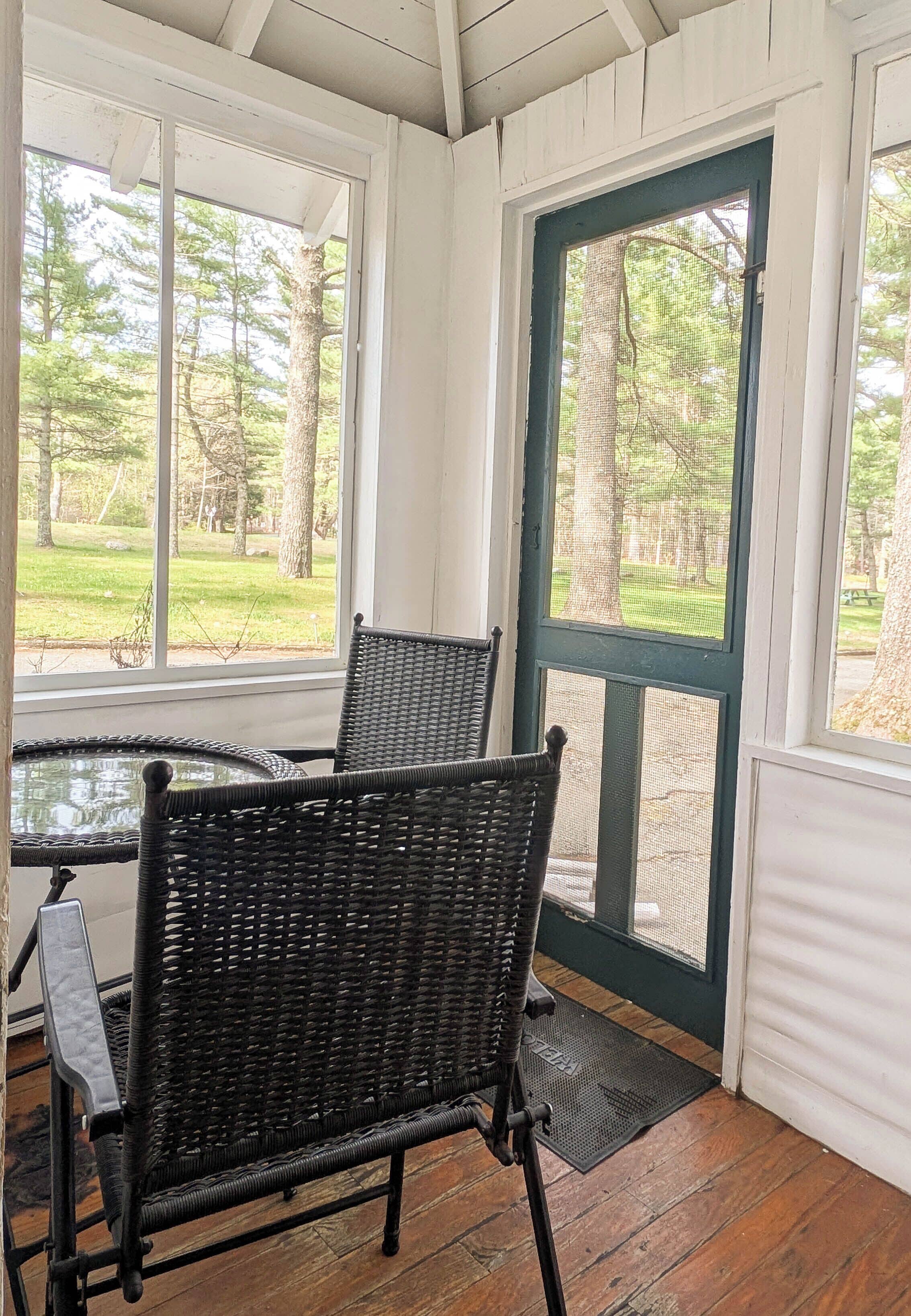 A covered porch with two wicker chairs and a small table, overlooking a grassy area with trees.