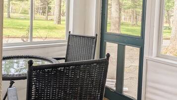 A covered porch with two wicker chairs and a small table, overlooking a grassy area with trees.