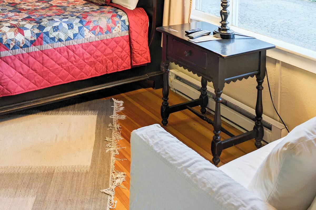A cozy bedroom with a wooden ceiling, a bed with a quilt, a bedside table with a lamp, and a window with curtains.