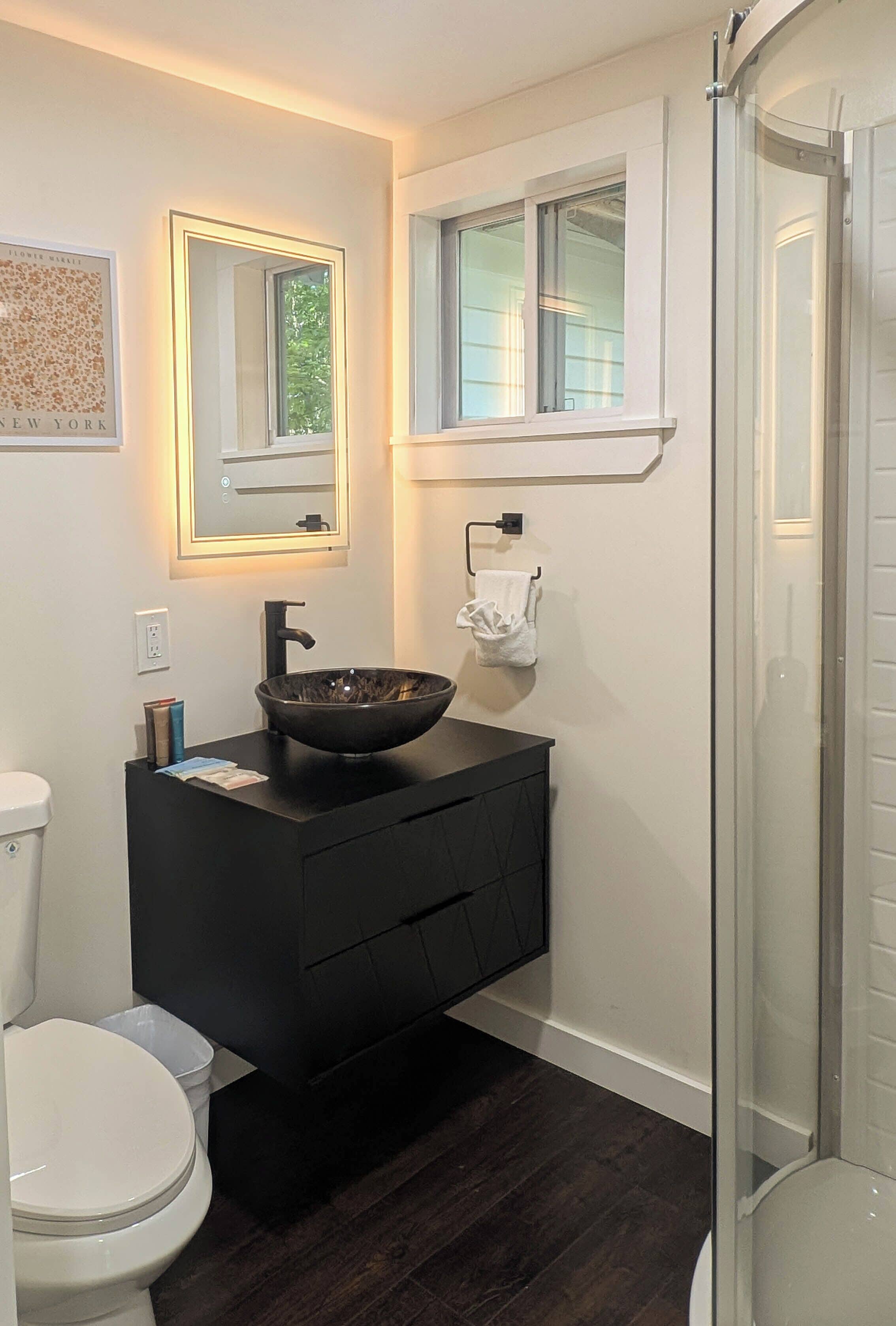 Un baño moderno con un tocador negro, un espejo grande, un lavabo tipo recipiente y una cabina de ducha con paredes de vidrio.
