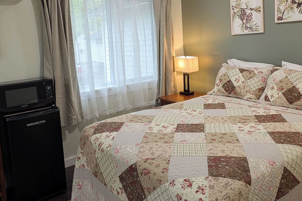 A bedroom with a bed, a nightstand, a lamp, a microwave, and a window with curtains.
