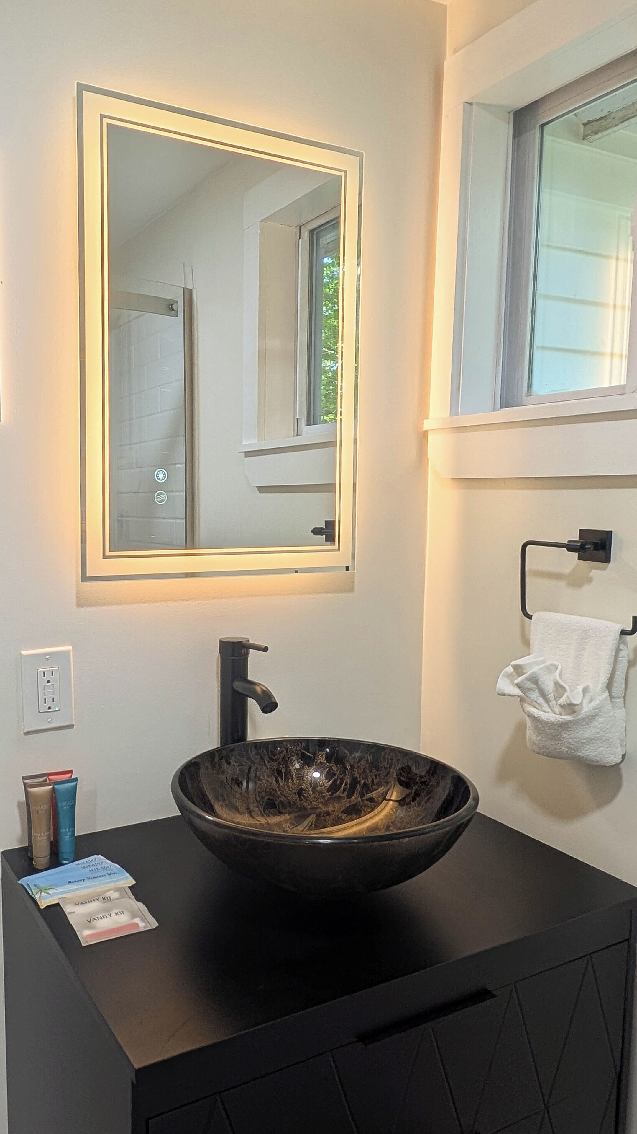 Een moderne badkamer met een zwart badkamermeubel, een grote spiegel met verlichting onder de rand, een zwart wasbak en een handdoekhouder met een witte handdoek.