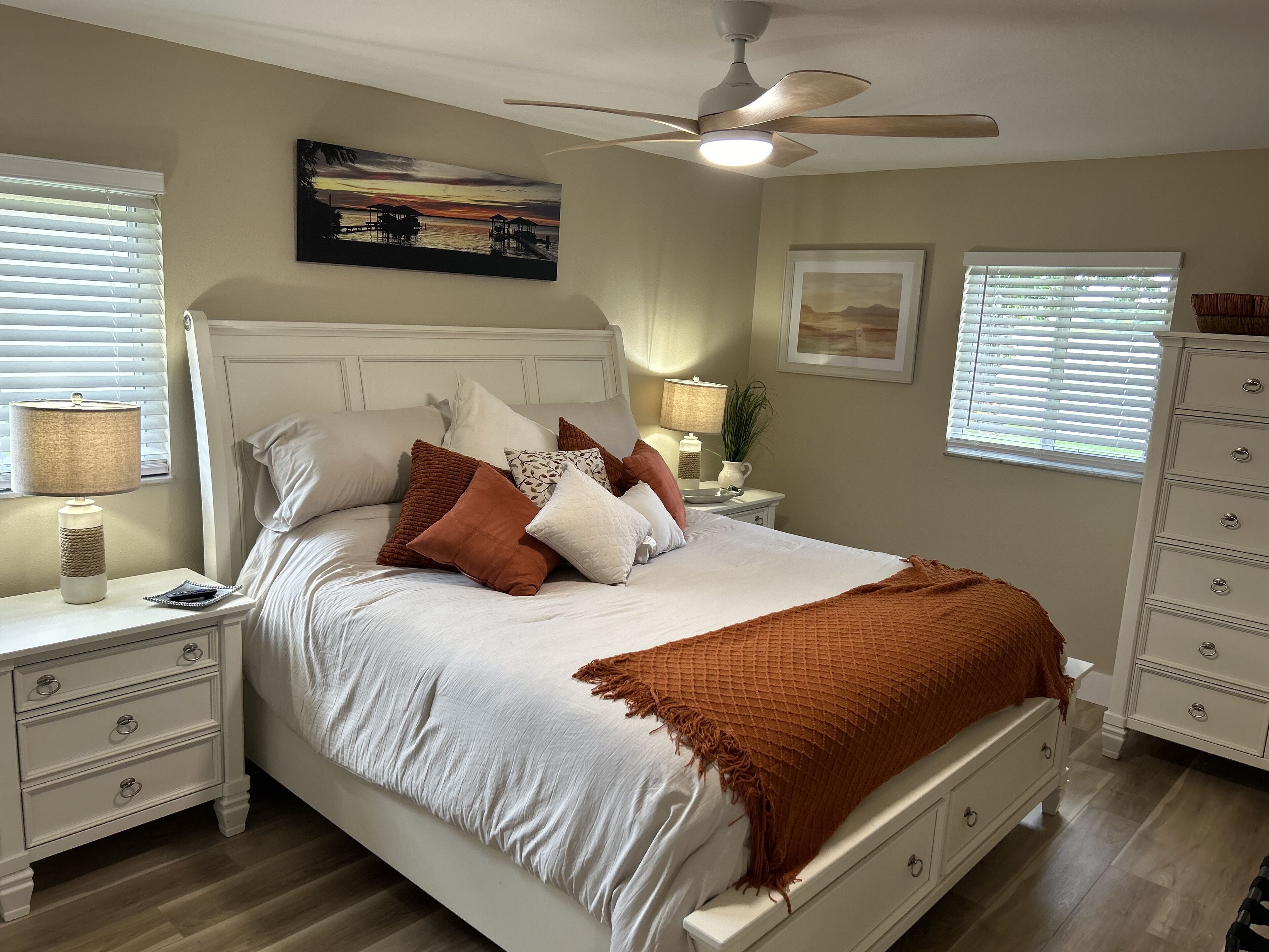 A neatly made bed with a white comforter and brown throw blanket, a ceiling fan, and artwork on the walls.