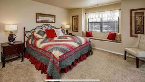 A bedroom with a large bed, a nightstand, a chair, a window with blinds, and framed artwork on the walls.