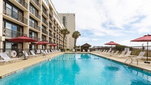 Una piscina amb gandules i ombrel·les, envoltada d'un edifici d'àmbit elevat amb balcons.