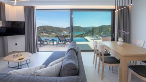 Ein modernes Wohnzimmer mit einer Couch, einem Esstisch und Stühlen, einem Flachbildfernseher und einer großen Schiebeglastür, die zu einem Pool und einem schönen Ausblick führt.