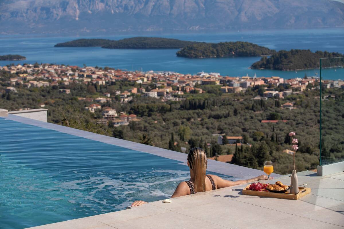 Una persona descansando en una piscina infinita con una vista panorámica de un pueblo costero y montañas al fondo.