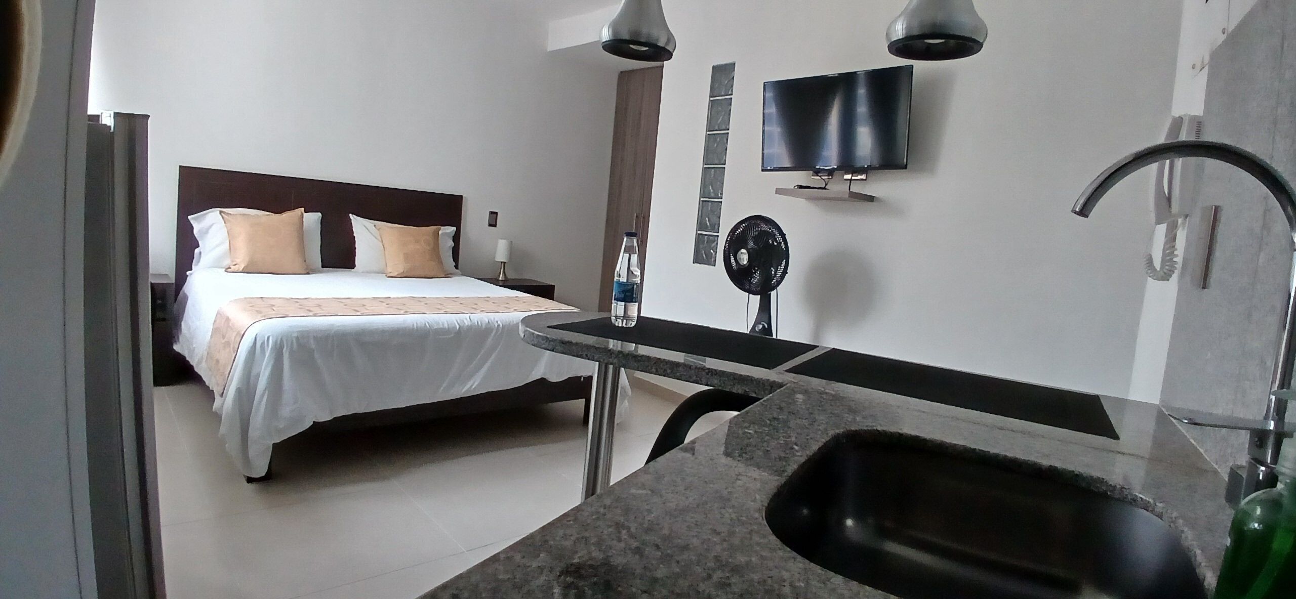 Ein Hotelzimmer mit einem Bett, einem an der Wand befestigten Fernseher, einem Ventilator und einem Waschbecken.
