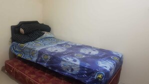 Quarto pequeno com uma cama de solteiro, colcha estampada e mala vermelha no chão.