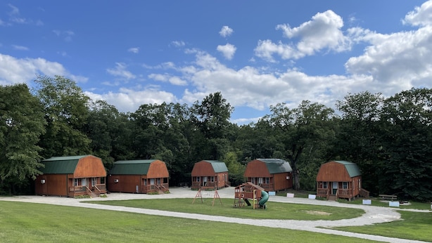 A row of wooden cabins with green roofs, surrounded by a grassy field and dense trees. - Sleeping Cabin #12 perfect to sleep up to 12 guests No Water in Cabin (Letts)