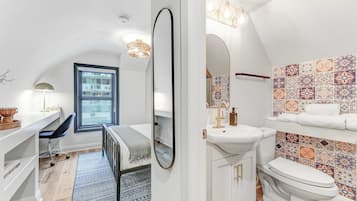 Un baño moderno con un tocador blanco, un espejo grande, un lavamanos y un inodoro. Hay una ventana con vistas al exterior, y las paredes están decoradas con baldosas estampadas.