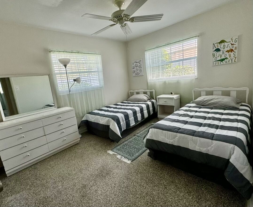 A bedroom with two beds, a dresser, a ceiling fan, and two windows with blinds.
