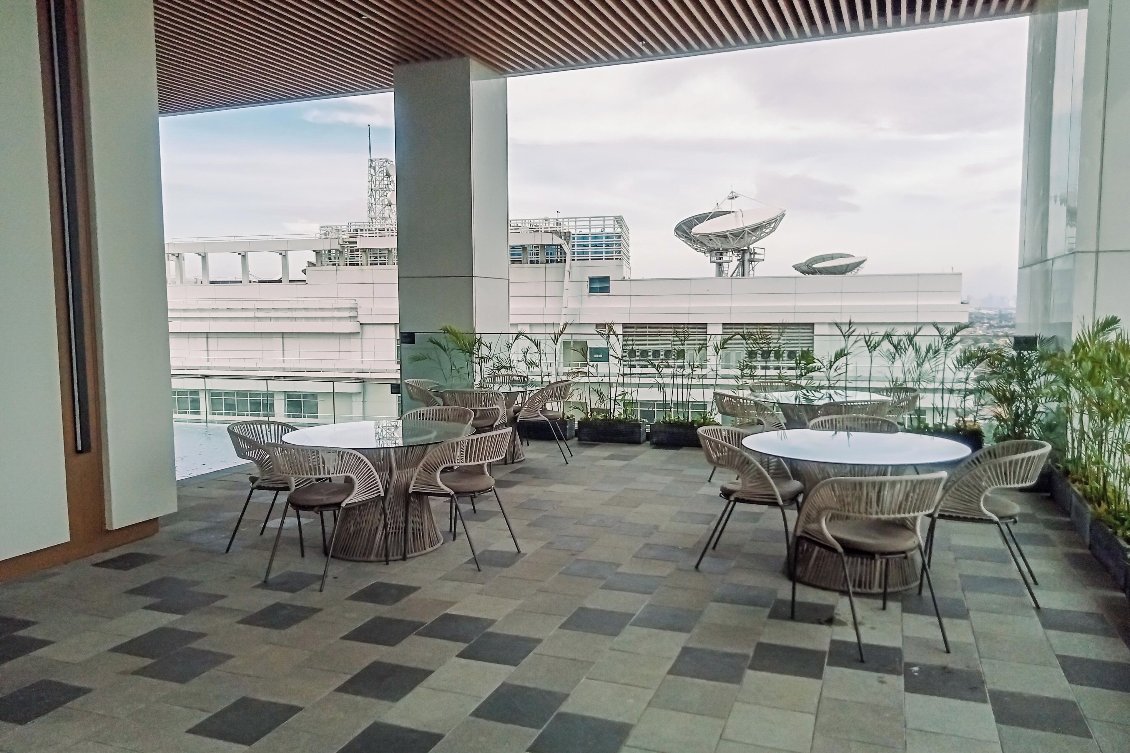 An outdoor terrace with round tables and wicker chairs, featuring a view of modern buildings and a satellite dish.