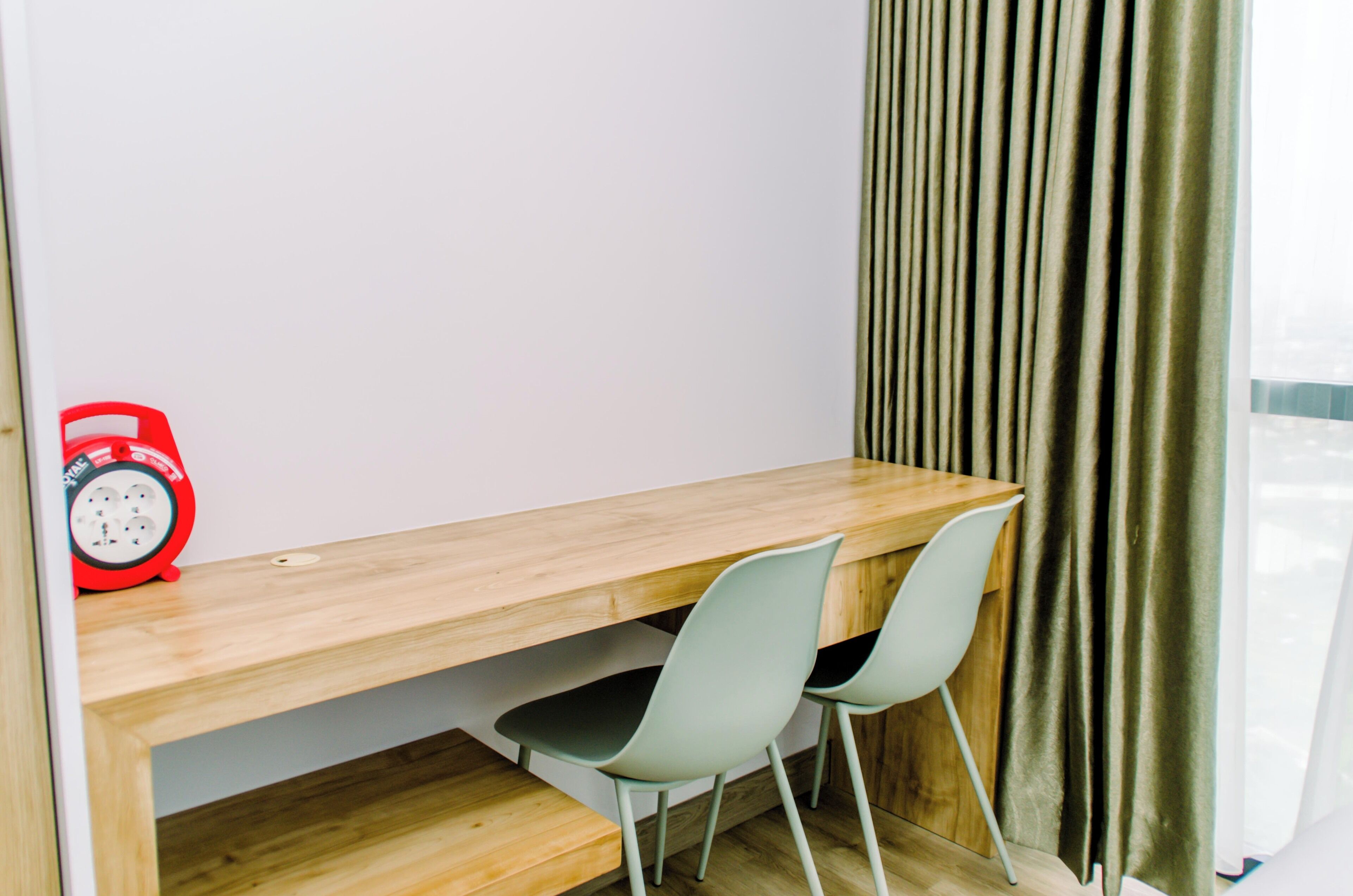 Une pièce avec un bureau en bois, deux chaises, une horloge rouge et une fenêtre avec des rideaux.
