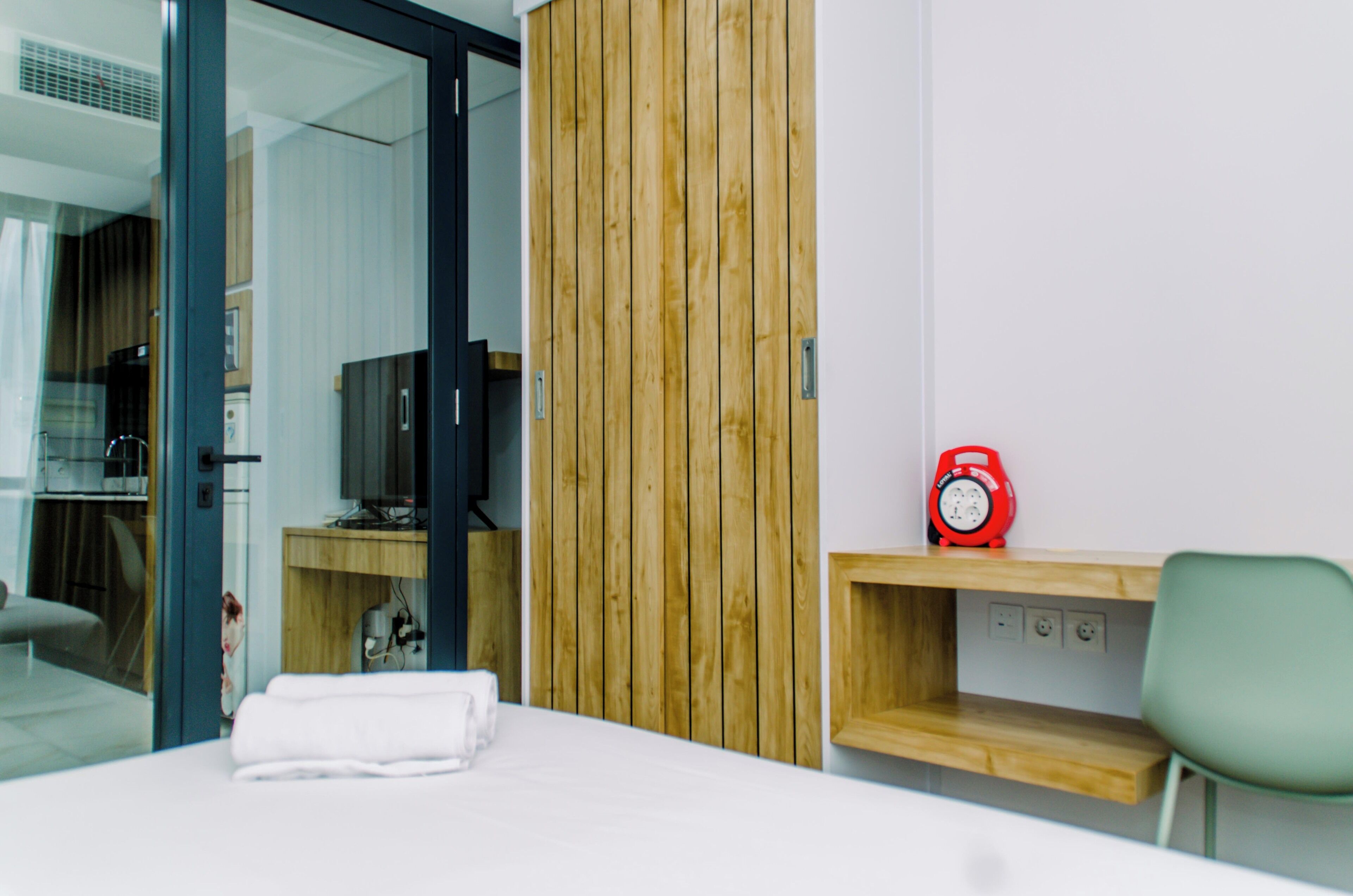 A hotel room with a bed, a wooden panel wall, a TV, a red alarm clock, and a green chair.