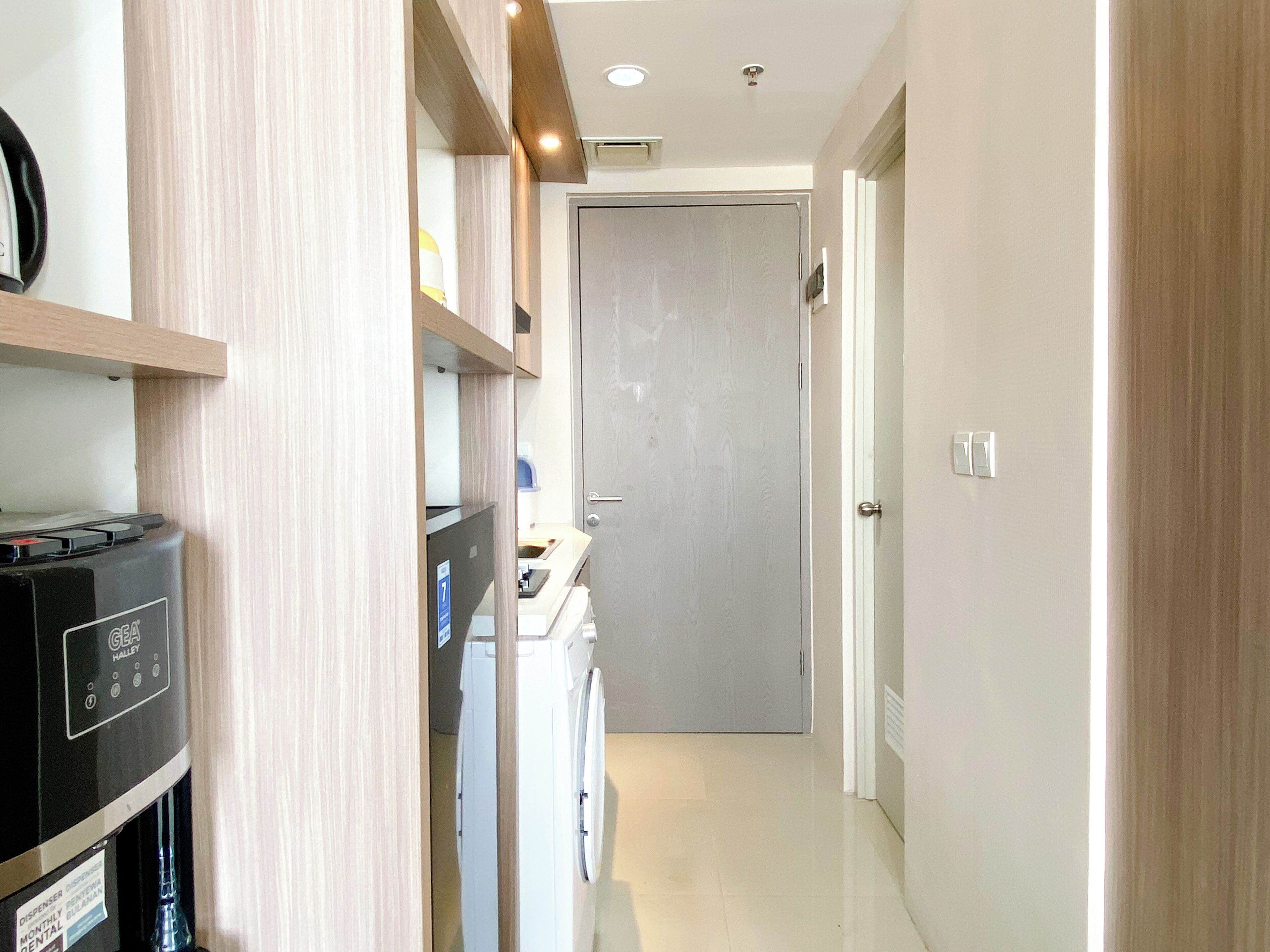 Un pasillo estrecho con lavadora, una pequeña cocina americana y una puerta que da a otra habitación.