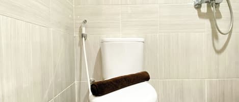 Inodoro blanco con una toalla marrón sobre el asiento, un cabezal de ducha manual y un dispensador de papel higiénico.