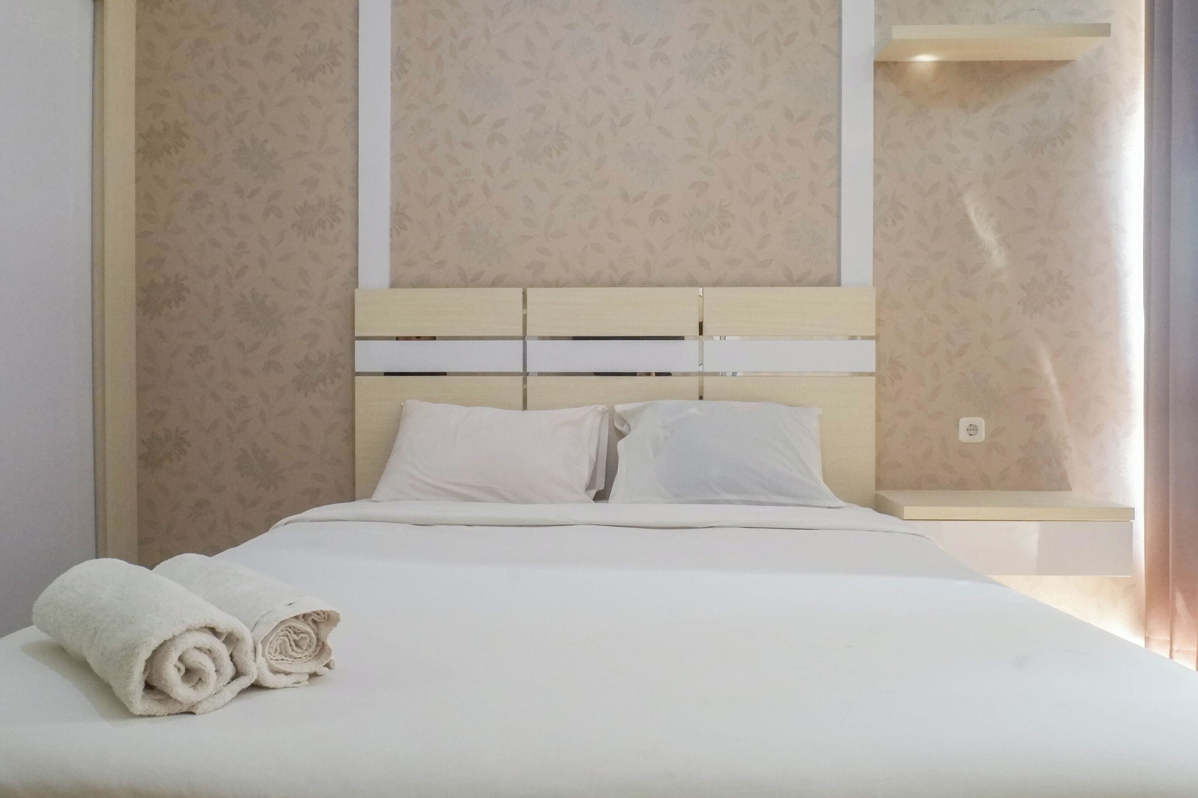 A neatly made bed with white linens and pillows, a rolled towel on the side, and a headboard with a light wood finish.