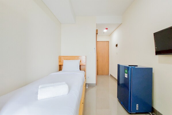 A hotel room with a single bed, a wooden headboard, a white pillow, a blue refrigerator, and a television mounted on the wall. - Cozy Studio (No Kitchen) Apartment B Residence (Pagedangan)