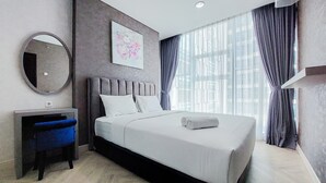 A modern hotel room with a large bed, a desk, a chair, a round mirror, and a large window with curtains. - Nice And Homey 1Br At Brooklyn Alam Sutera Apartment (South Tangerang)