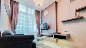 A modern living room with a sofa, a round coffee table, a TV mounted on the wall, and a large window with curtains. - Nice And Homey 1Br At Brooklyn Alam Sutera Apartment (South Tangerang)