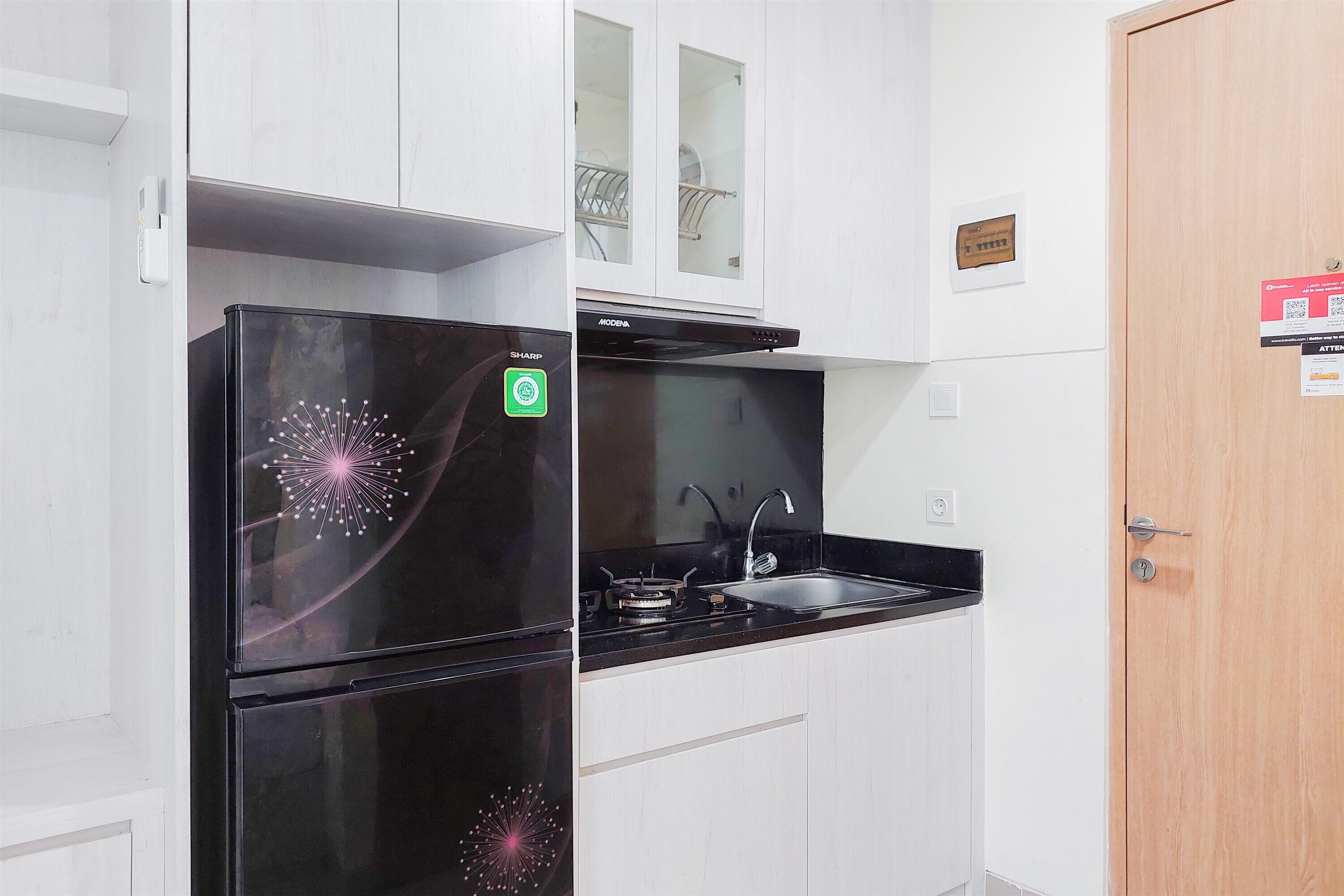 Une cuisine de taille réduite avec un réfrigérateur noir, une cuisinière et un évier. Une porte en bois se trouve à droite et un meuble blanc est placé au-dessus de l’évier.