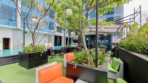 Un área de estar al aire libre con mobiliario de mimbre y macetas, una piscina y un edificio moderno al fondo.