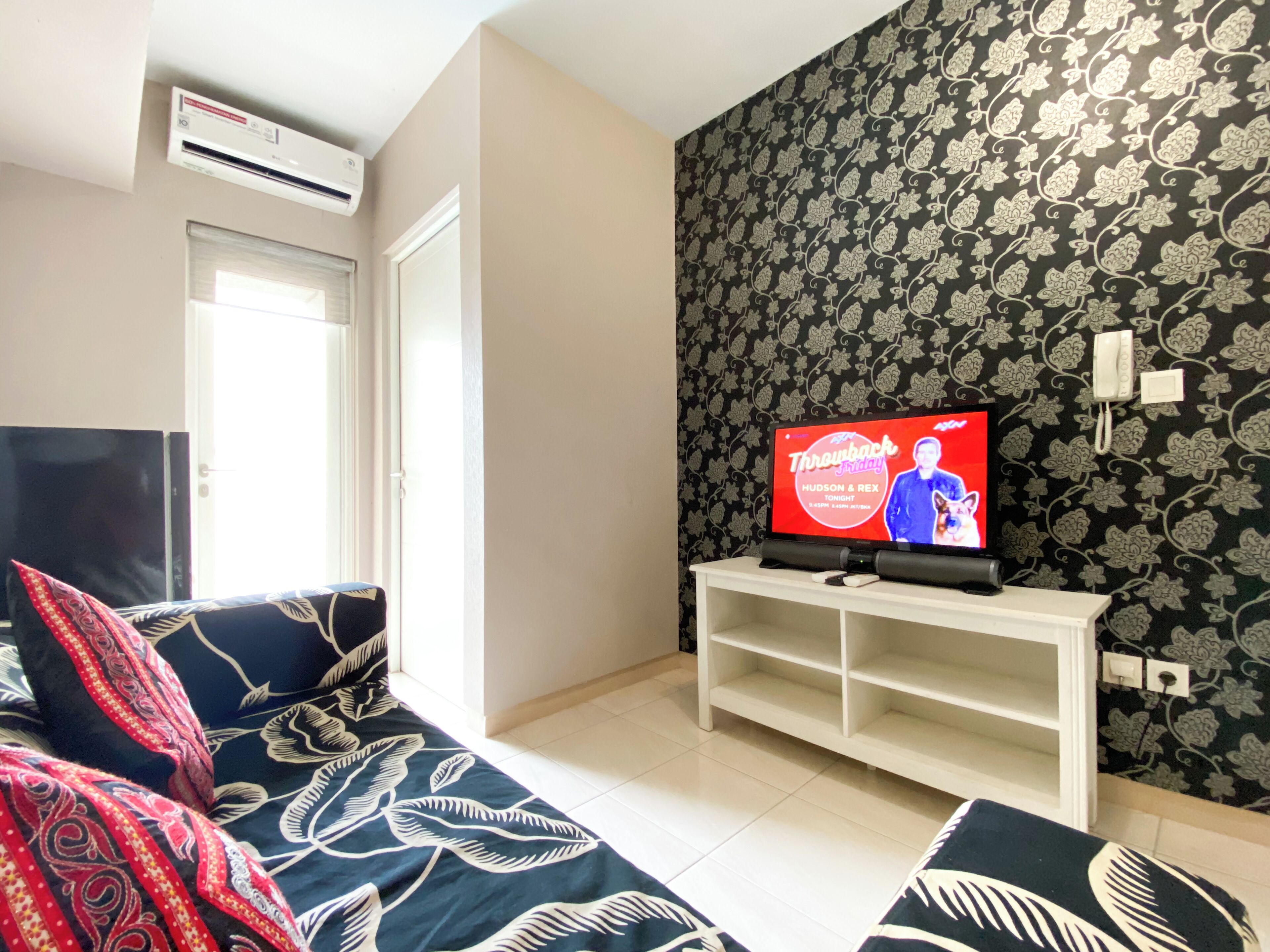 A living room with a sofa, a TV on a stand, a patterned wall, and an air conditioning unit.