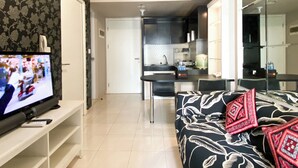 Ein modernes Wohnzimmer mit Flachbildfernseher, einer Couch mit gemusterten Kissen, einem Essbereich mit Tisch und Stühlen sowie einer Küche mit dunklen Schränken und einer gefliesten Spritzschutzwand.